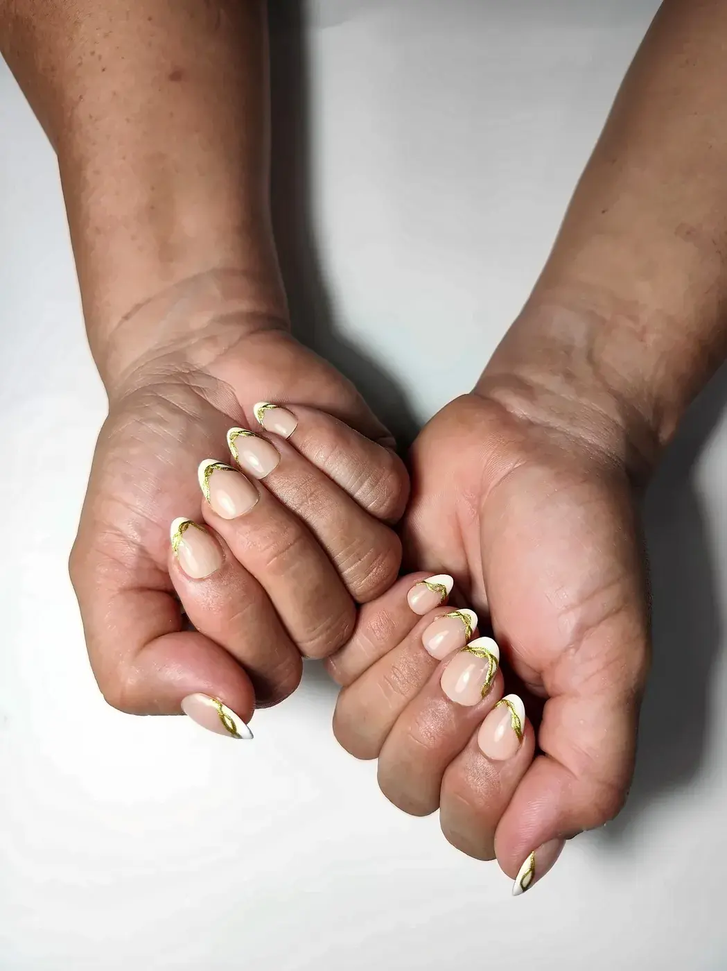Manos entrelazadas con uñas postizas almendra cortas en nude con francesa blanca y línea dorada ondulada