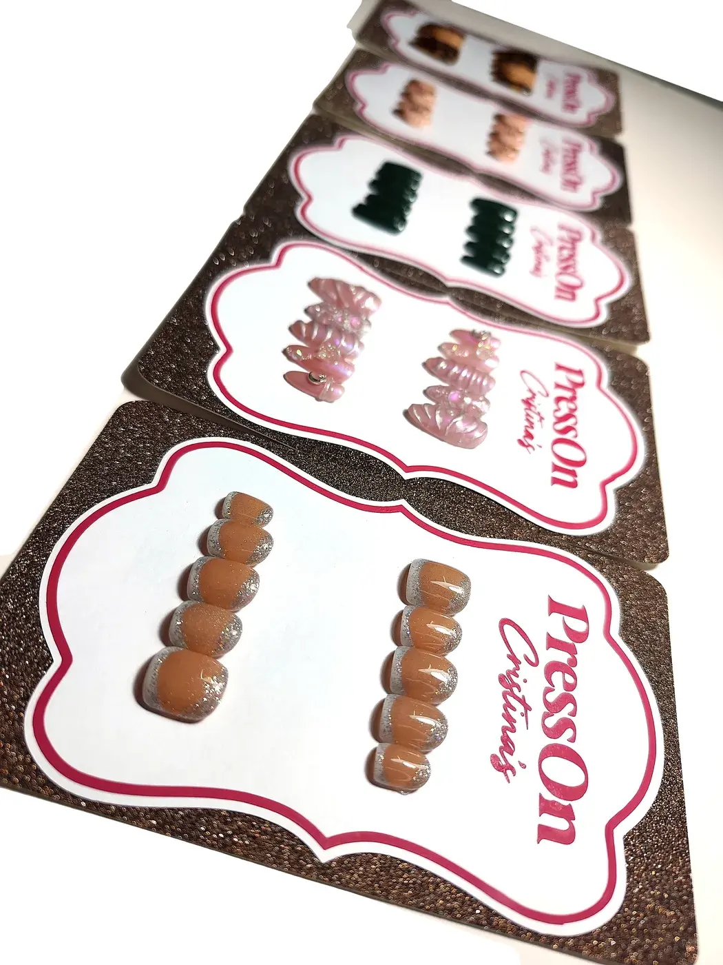 Varias cartulinas PressOn Cristina's en fila con sets distintos: nude con francesa plateada, rosa perlado 3D, verde oscuro y carey