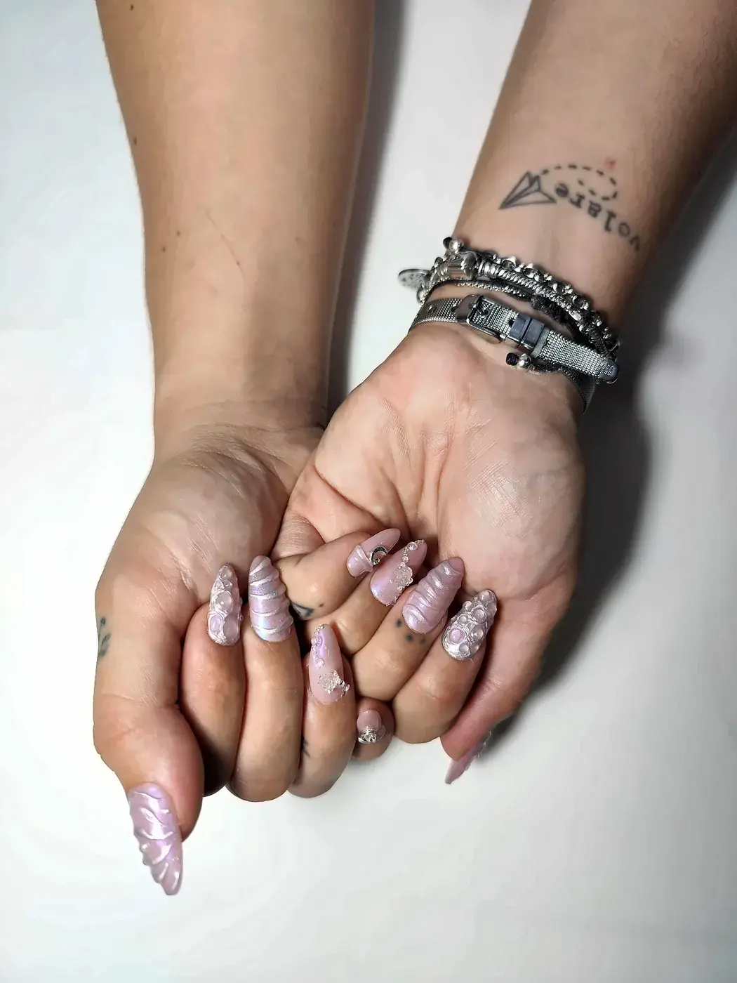 Manos juntas con uñas postizas almendra largas en rosa nacarado con relieve 3D, efecto cromado unicornio, perlas y charms de luna y rosa