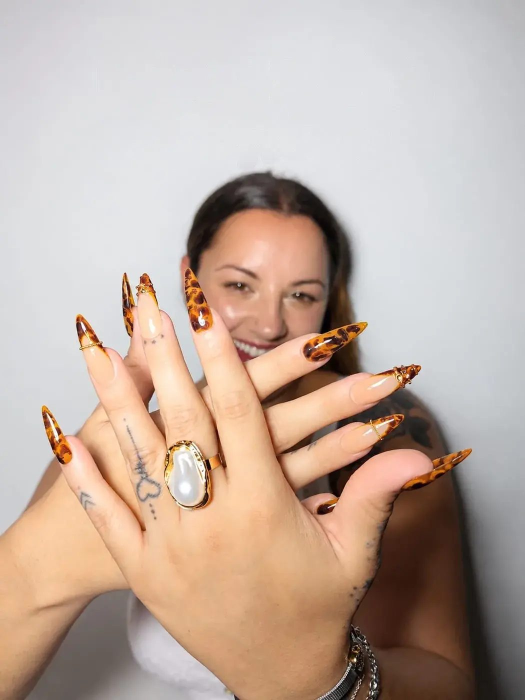 Cristina sonriendo detrás de sus manos con uñas postizas stiletto largas de carey, acentos nude con charms dorados y anillo de perla